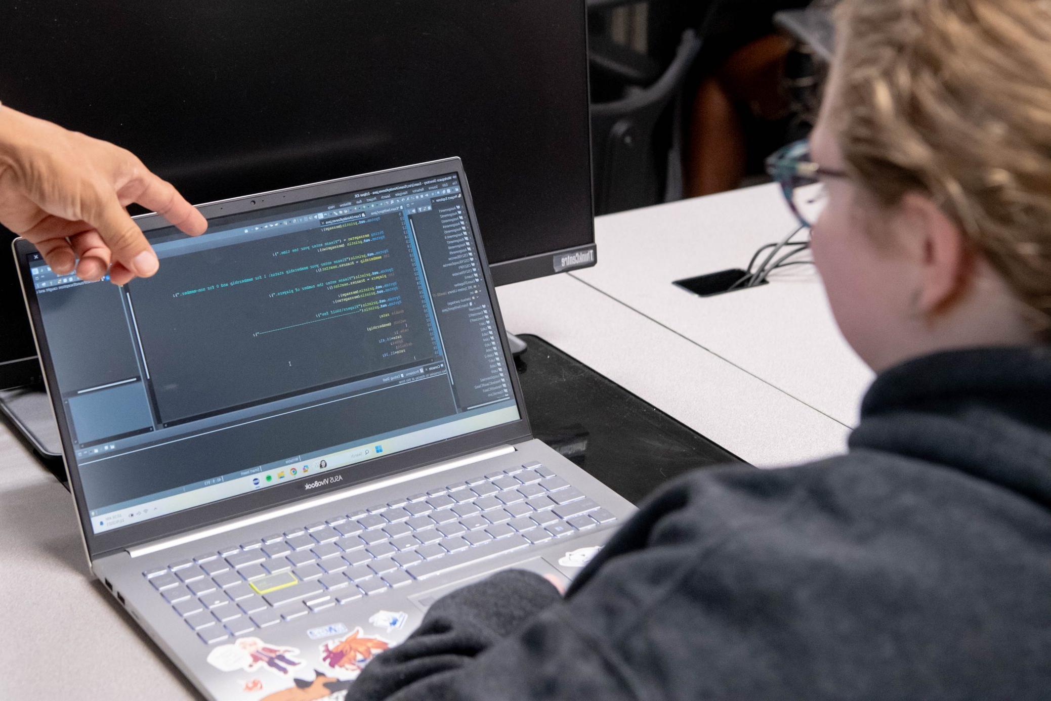 Student sitting in front of laptop with professors finger pointing at code on computer screen.